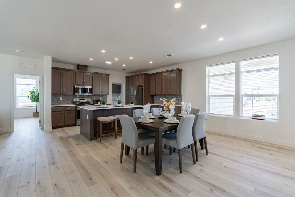 Modern kitchen with wooden cabinets and island, adjacent to a dining area with a dark table and four chairs. Large windows and recessed lighting illuminate the space with light wood flooring.