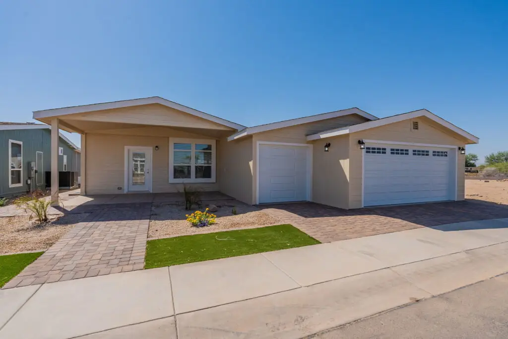 Single-story house with two-car garage, light beige exterior, paved driveway, and desert landscaping under a clear blue sky.