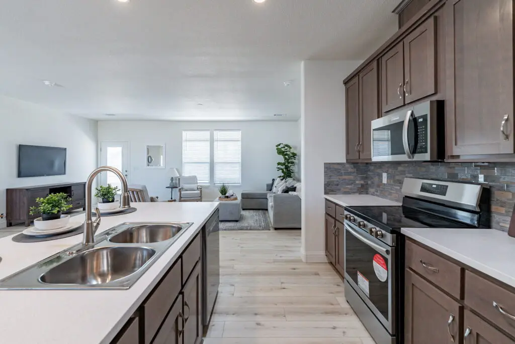 Modern kitchen with stainless steel appliances, a double sink, and dark wood cabinets. The kitchen opens into a living area with a sofa, TV, and large windows.