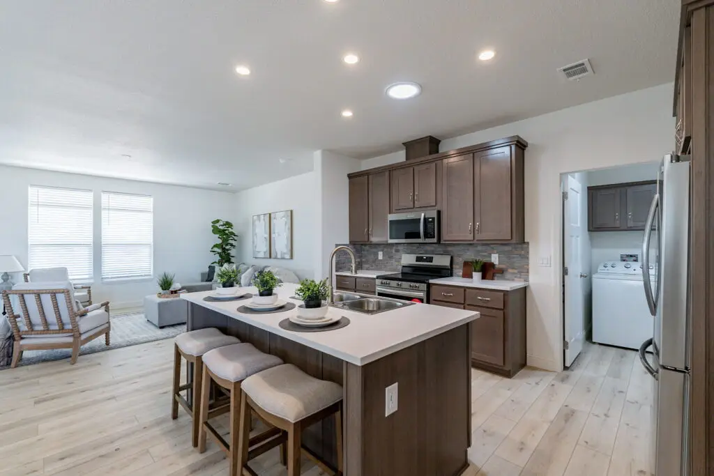 Modern kitchen with an island, three stools, stainless steel appliances, and adjacent laundry room. Living area with plants and seating is visible in the background.