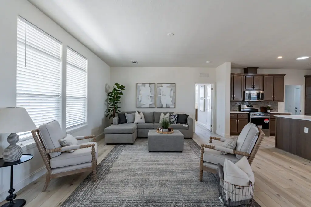 A modern living room with a gray sofa, armchairs, and a coffee table on a patterned rug. There are artworks above the sofa and a plant in the corner. A kitchen is visible in the background.