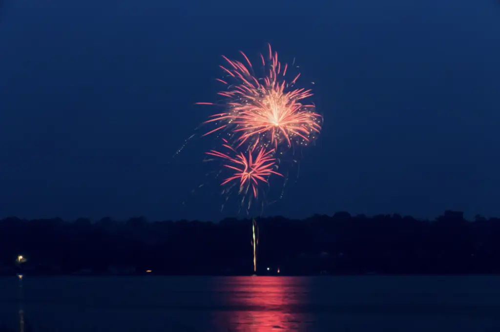The fireworks display above Lake Conroe casts vibrant pink and red bursts, their colors dancing across the dark water, creating a dazzling spectacle perfect for the holidays.
