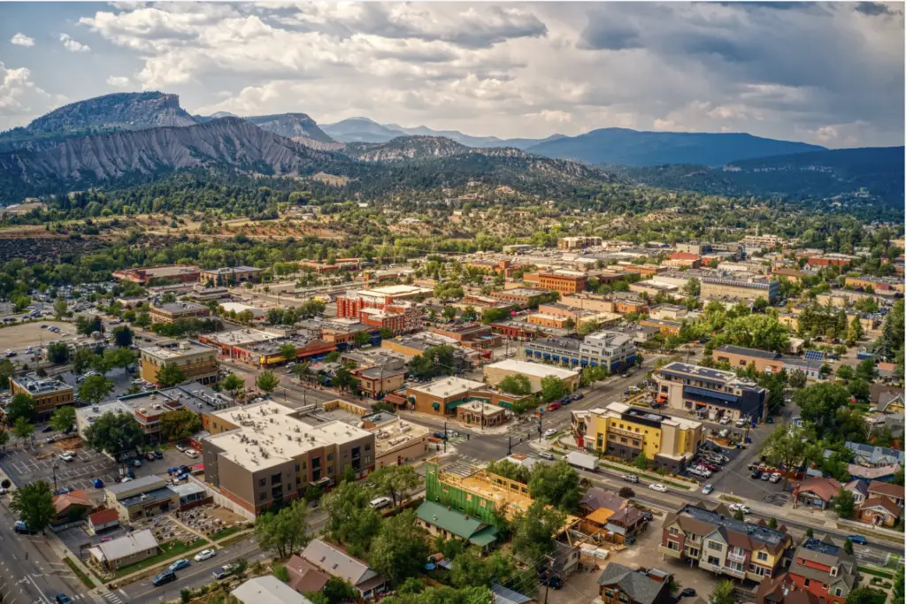Aerial view of a small town with buildings, roads, and vehicles, surrounded by trees and mountains under a partly cloudy sky—truly the perfect time for an outdoor adventure.
