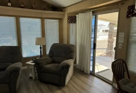 This cozy living room, featuring two gray recliners, a lamp, and a small table, is nestled within the charming 1993 Cavco on Lot 482. A sliding glass door with curtains opens up to an inviting outdoor area.