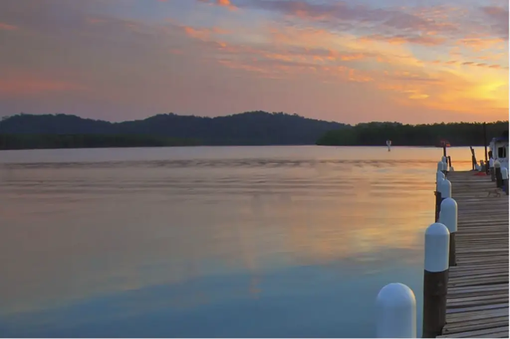 A serene lakeside retreat at sunset features a wooden dock extending into the water. The sky displays shades of orange, pink, and purple, reflecting on the calm surface—a perfect Thanksgiving escape with hills visible on the horizon.