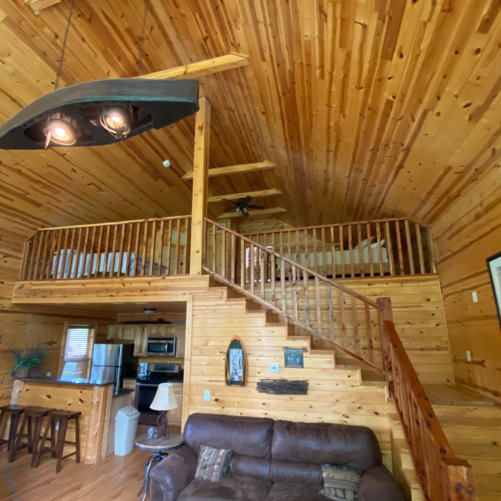 The charming log cabin interior, perfect for a Spring Break getaway, boasts wooden walls and ceilings. It features a loft with a railing, stairs, a comfortable leather sofa, and a kitchen area with bar stools. Just steps away from the serene beauty of Lake Conroe.