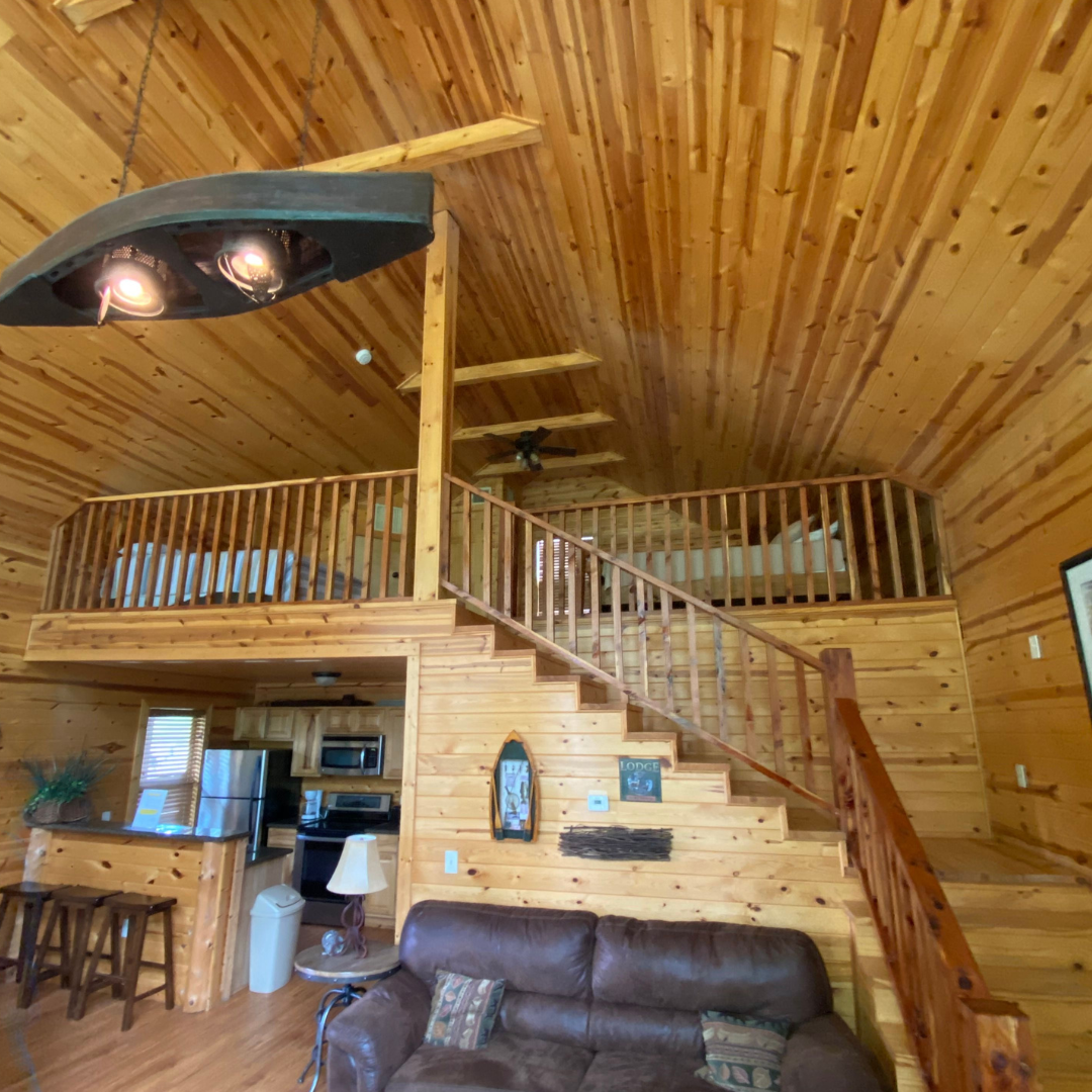 The charming log cabin interior, perfect for a Spring Break getaway, boasts wooden walls and ceilings. It features a loft with a railing, stairs, a comfortable leather sofa, and a kitchen area with bar stools. Just steps away from the serene beauty of Lake Conroe.