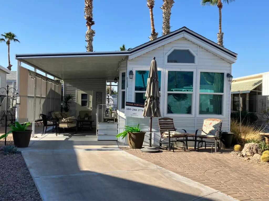A small, white 1993 Skyline mobile home with a covered patio, outdoor furniture, potted plants, and a "home for sale" sign graces Lot 291. Palm trees sway under the clear sky in the background.