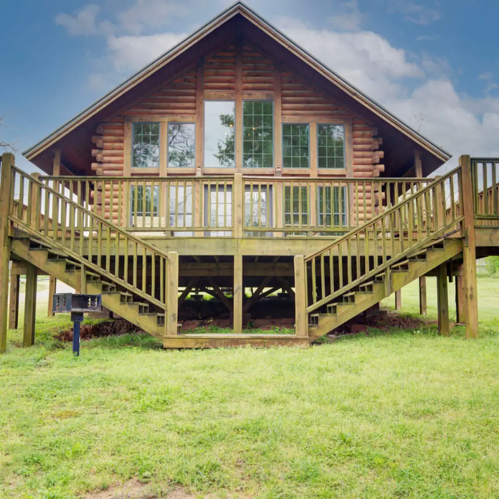 Nestled at the water's edge of Lake Conroe, this wooden cabin boasts a triangular roof and large windows, surrounded by a wooden deck and steps on a grassy area—perfect for family fun.