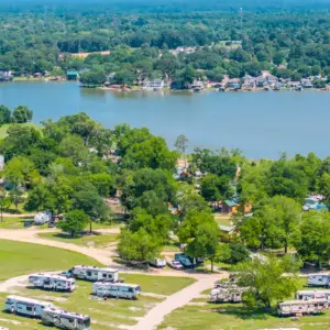 Aerial view of a campground with RVs nestled among trees at the water's edge, perfect for long-term RV stays. In the background, houses are scattered throughout a lush forested area near the tranquil lake.