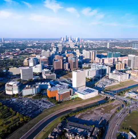 Aerial view of a large cityscape with tall buildings, roads, and patches of greenery under a clear blue sky. Nestled within the urban sprawl is Rayford Crossing, offering comfort and convenience to medical center visitors navigating this expansive metropolis.