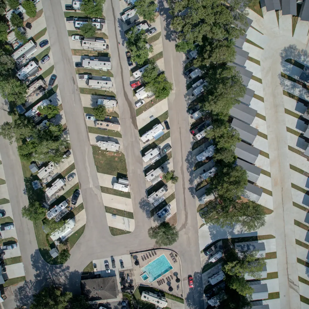 Aerial view of an RV park nestled among majestic pines, featuring rows of parked vehicles and covered RV spots. A small central swimming pool adds charm to this convenient stopover.
