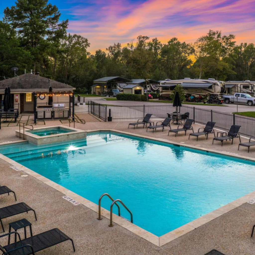 Outdoor swimming pool and hot tub surrounded by lounge chairs and a gazebo at the Majestic Pines RV park, with trees and RVs in the background during a stunning sunset. Perfect for a Spring Travel retreat, inviting you to stay longer and enjoy the serene atmosphere.
