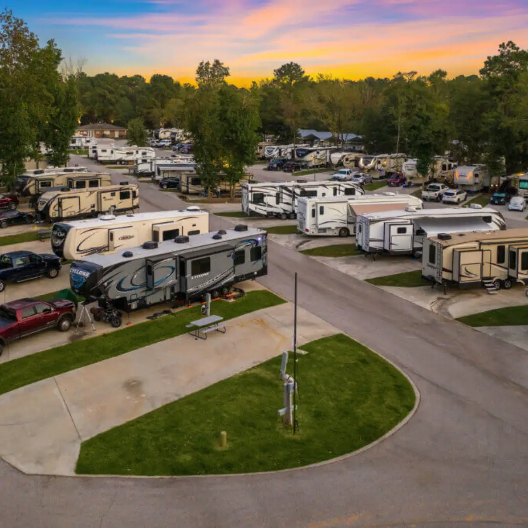 Aerial view of a campground with numerous parked RVs on paved sites, nestled among majestic pines, under a colorful sunset sky.