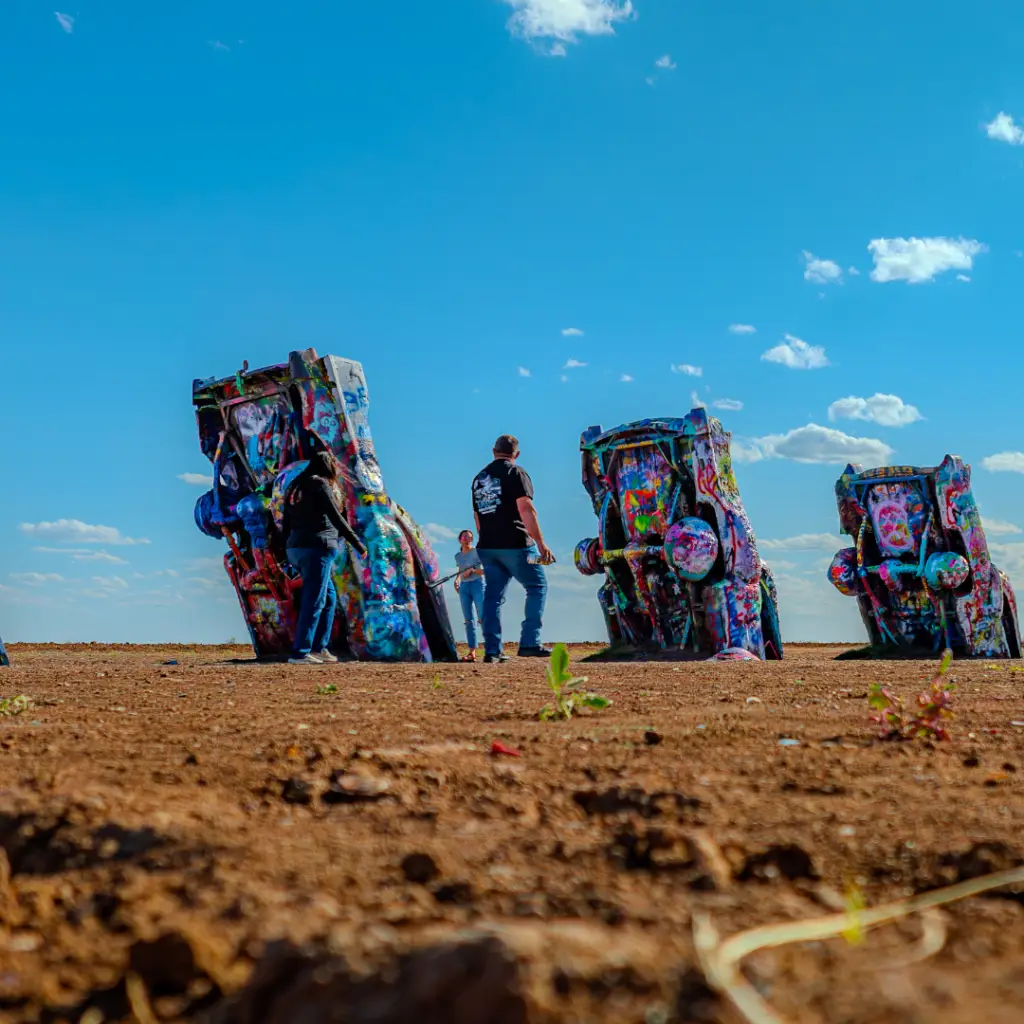 In Amarillo, people interact with half-buried, colorful painted cars under a clear blue sky in a desert-like area, creating lively events that capture the vibrant spirit of spring.