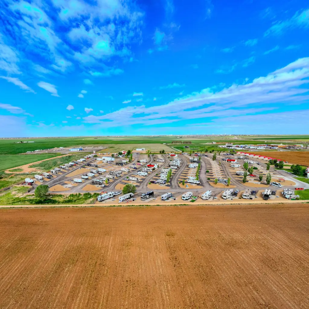 Aerial view of a large RV park nestled near the water's edge, surrounded by green fields under a clear blue sky with scattered clouds—an epitome of ultimate relaxation in the world of RV living.