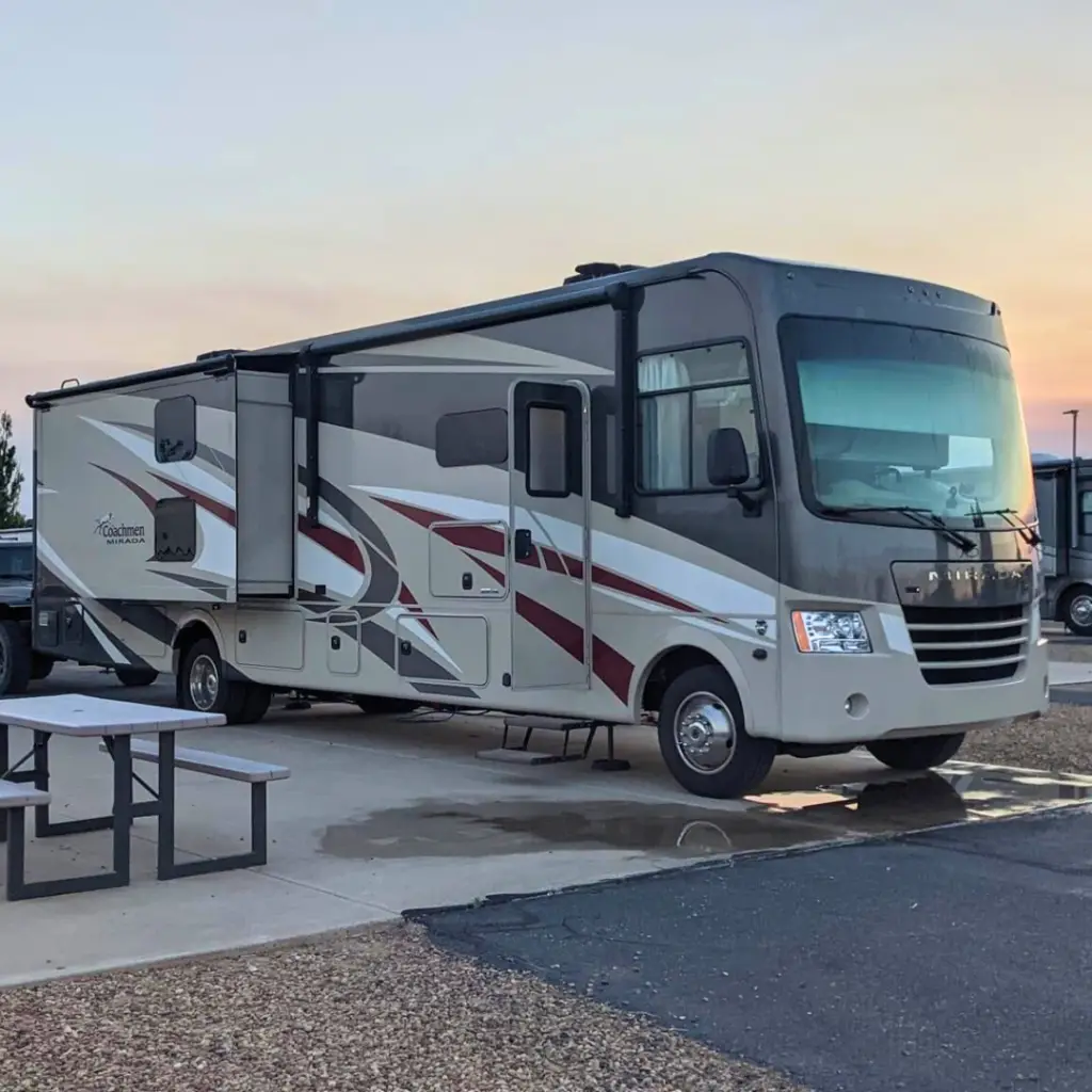 A large motorhome, ideal for both monthly RV rental and flexible RV options, is parked at a campsite with an extended awning. A picnic table sits nearby under the clear sky, inviting relaxation.