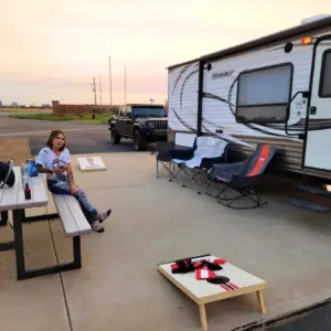 A person sits at a picnic table near a parked trailer and Jeep, enjoying the serene sunset. Nearby, two folding chairs and a cornhole game set the scene for relaxed camping vibes, perfect for those exploring monthly RV rental options.