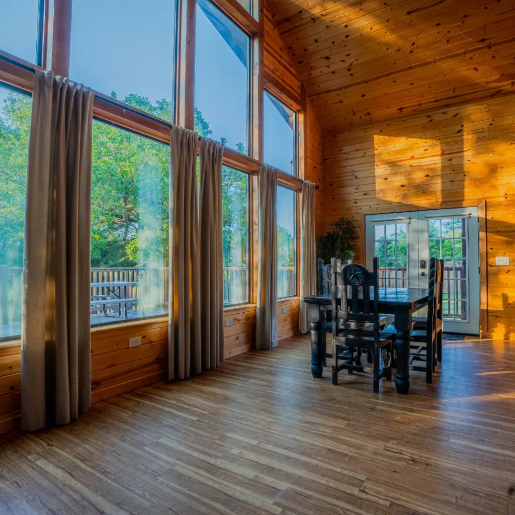 A sunlit room with large windows, wooden walls, and a wooden dining table with chairs offers a serene view. Curtains hang beside the windows, and a door opens to a deck outside, perfect for relaxing after enjoying water-based activities at nearby Lake Conroe.