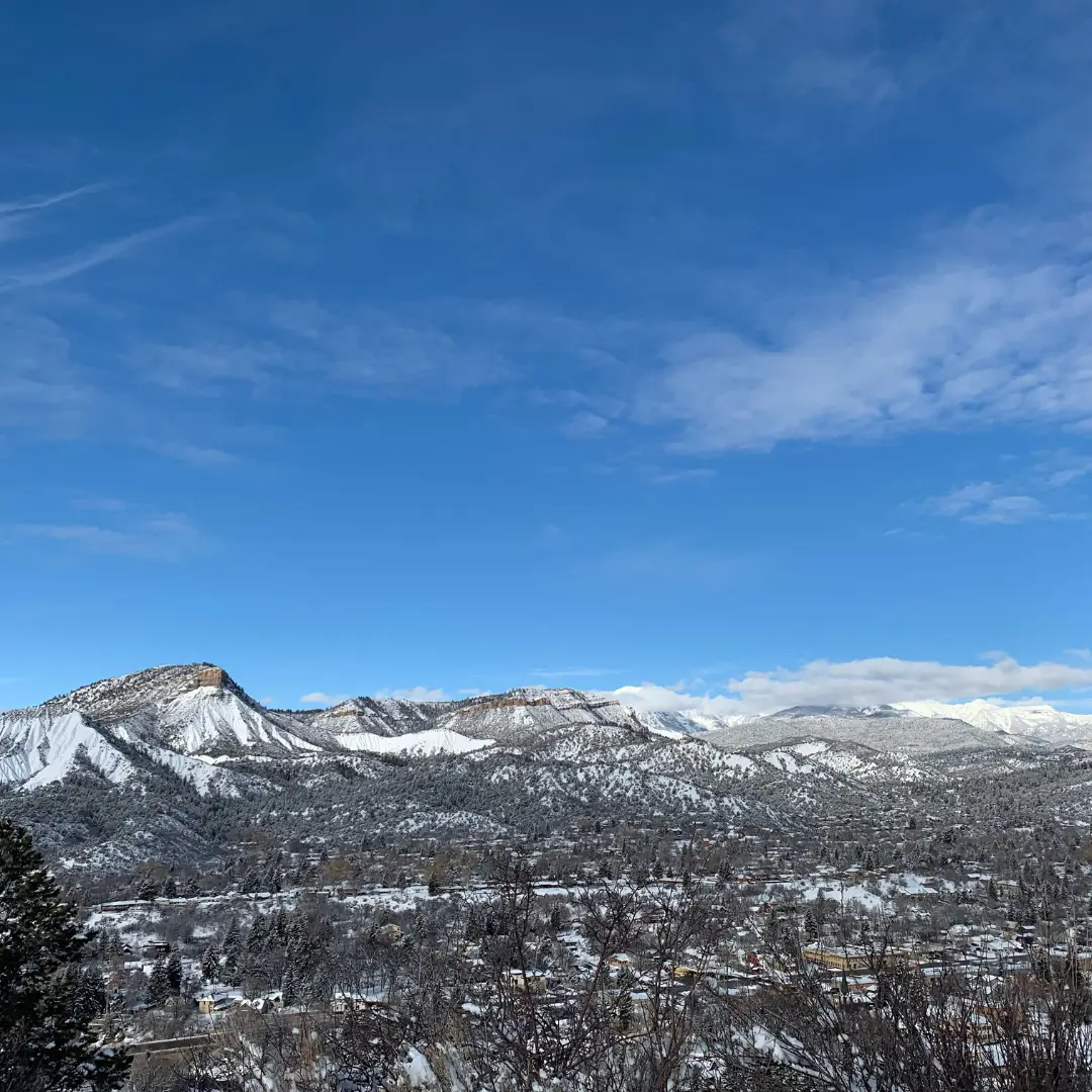 Snow-covered mountains surround the town of Durango under a clear blue sky dotted with scattered clouds. Bare trees stand in the foreground, inviting you to explore Durango's charming landscape and embrace spring activities amidst this picturesque setting.