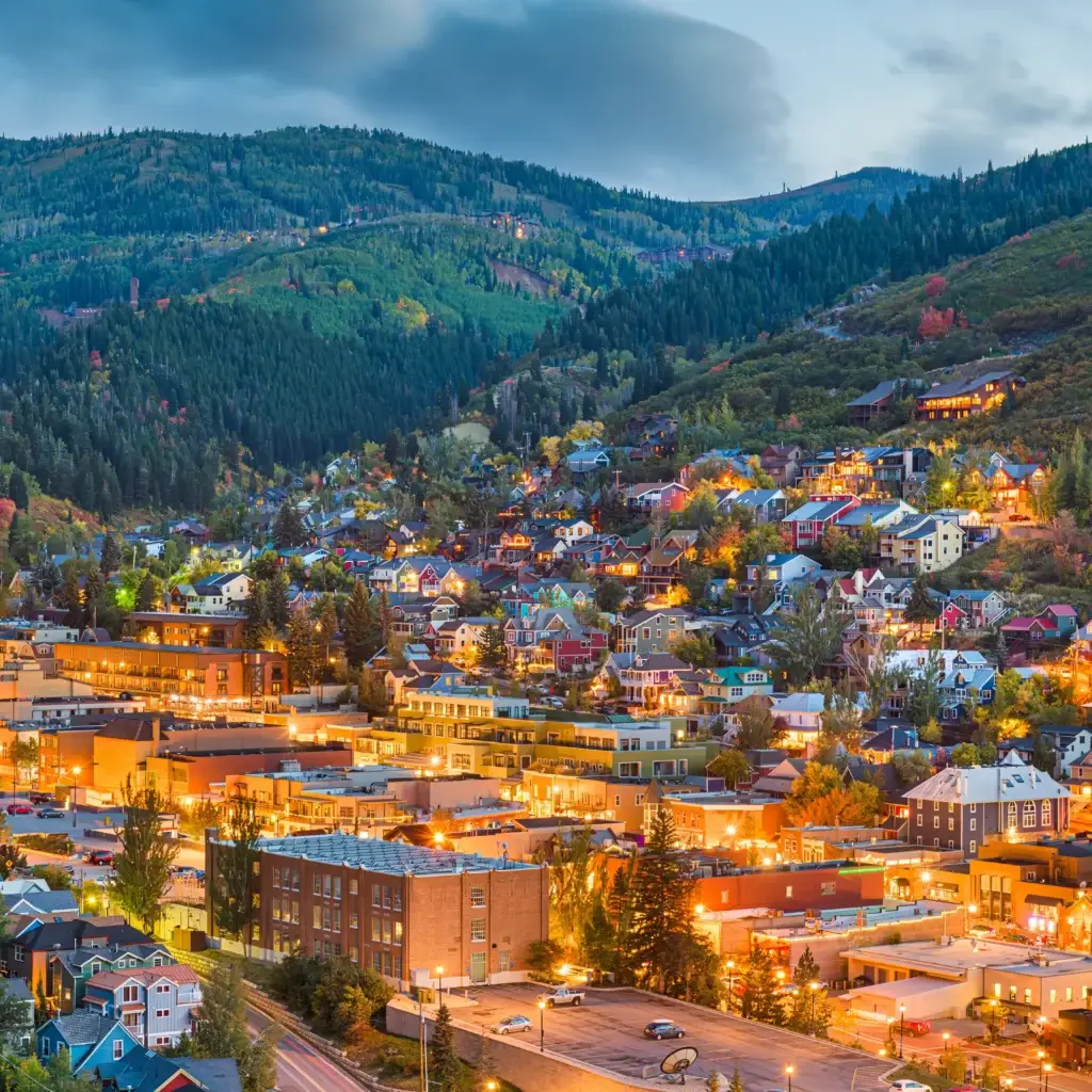 A scenic view of a mountainside town at dusk, with houses and buildings illuminated in Park City. Green hills and a cloudy sky form the perfect backdrop for your Summer RV Escape.