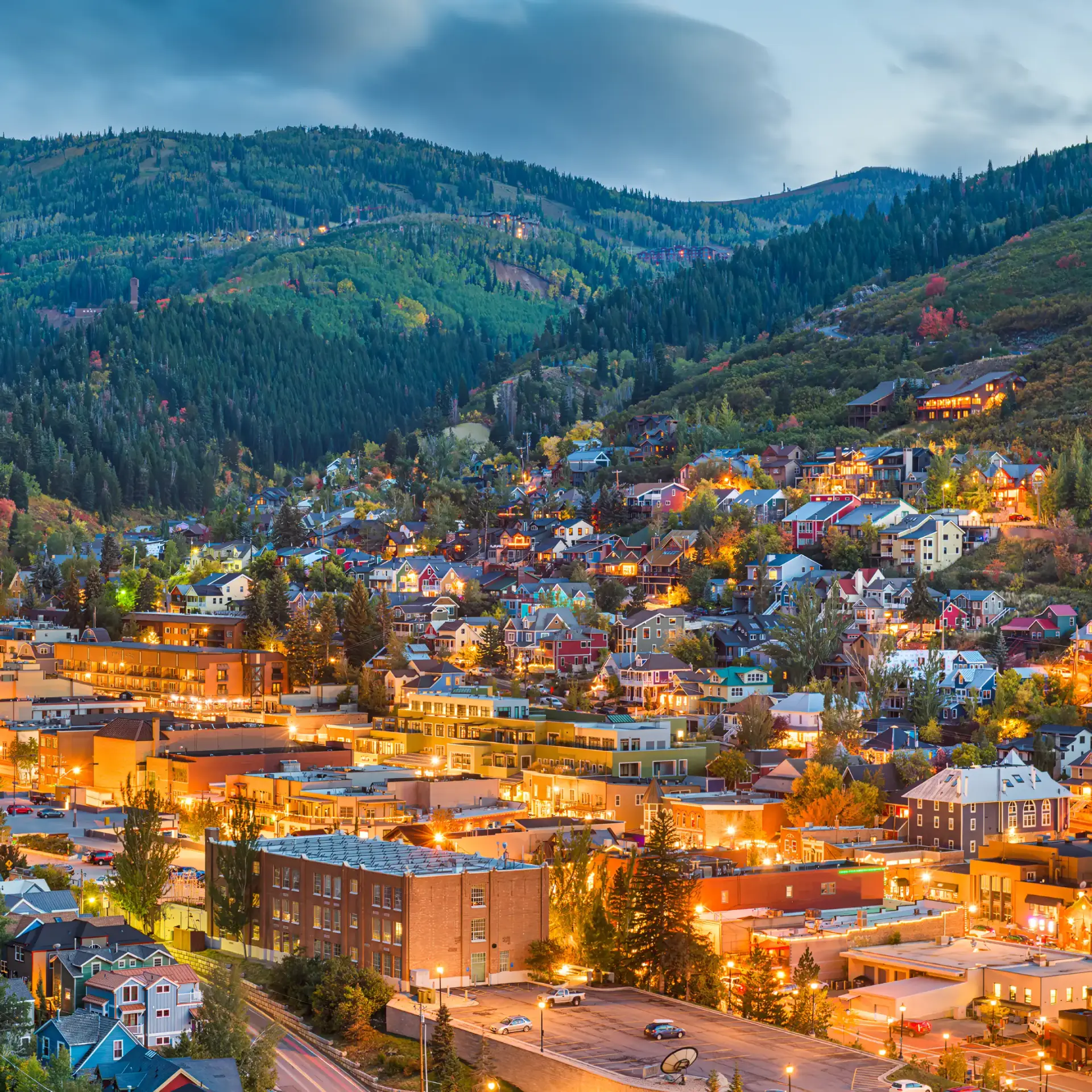 A scenic view of a mountainside town at dusk, with houses and buildings illuminated in Park City. Green hills and a cloudy sky form the perfect backdrop for your Summer RV Escape.