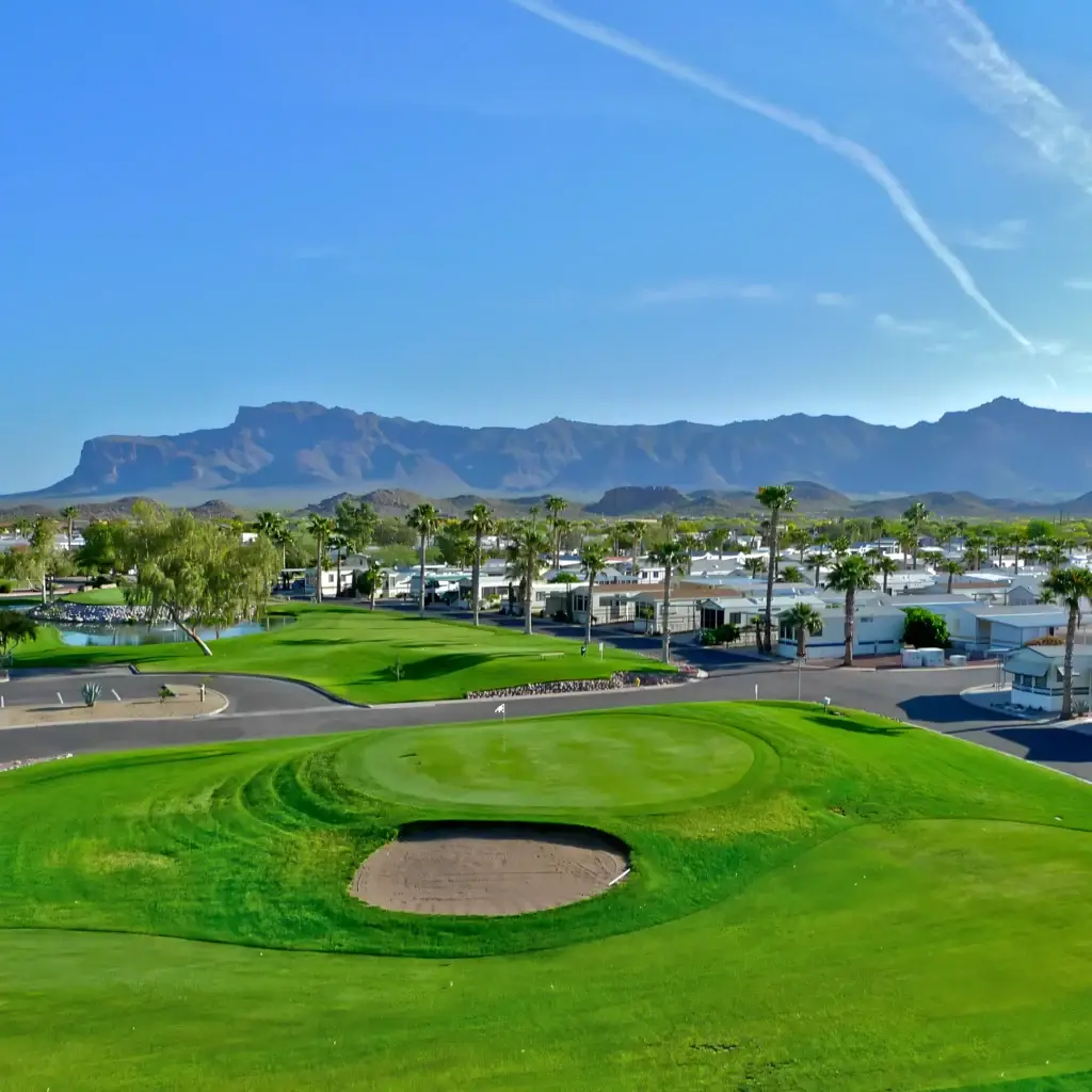 A golf course with a view of distant mountains under a clear blue sky, perfect for winter visitors. The area is surrounded by palm trees and low buildings, offering a secure RV site for those seeking both relaxation and peace.