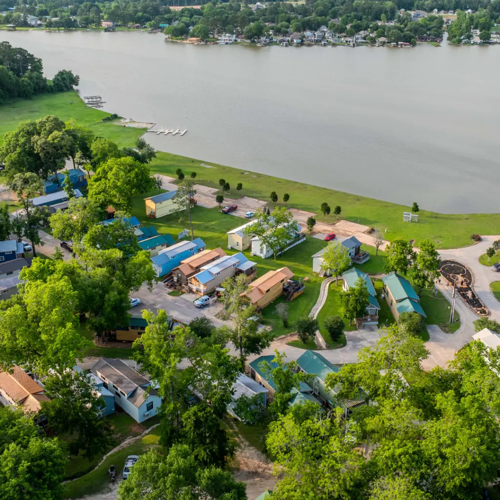 Aerial view of a serene lakeside community at the water's edge, where green trees surround blue-roofed buildings and a river with a dock and boats invites ultimate relaxation.