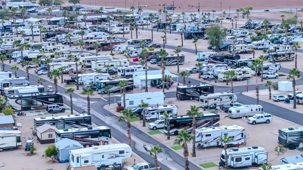 Rows of RVs are parked in a secure RV site within a desert campground, with palm trees scattered throughout.