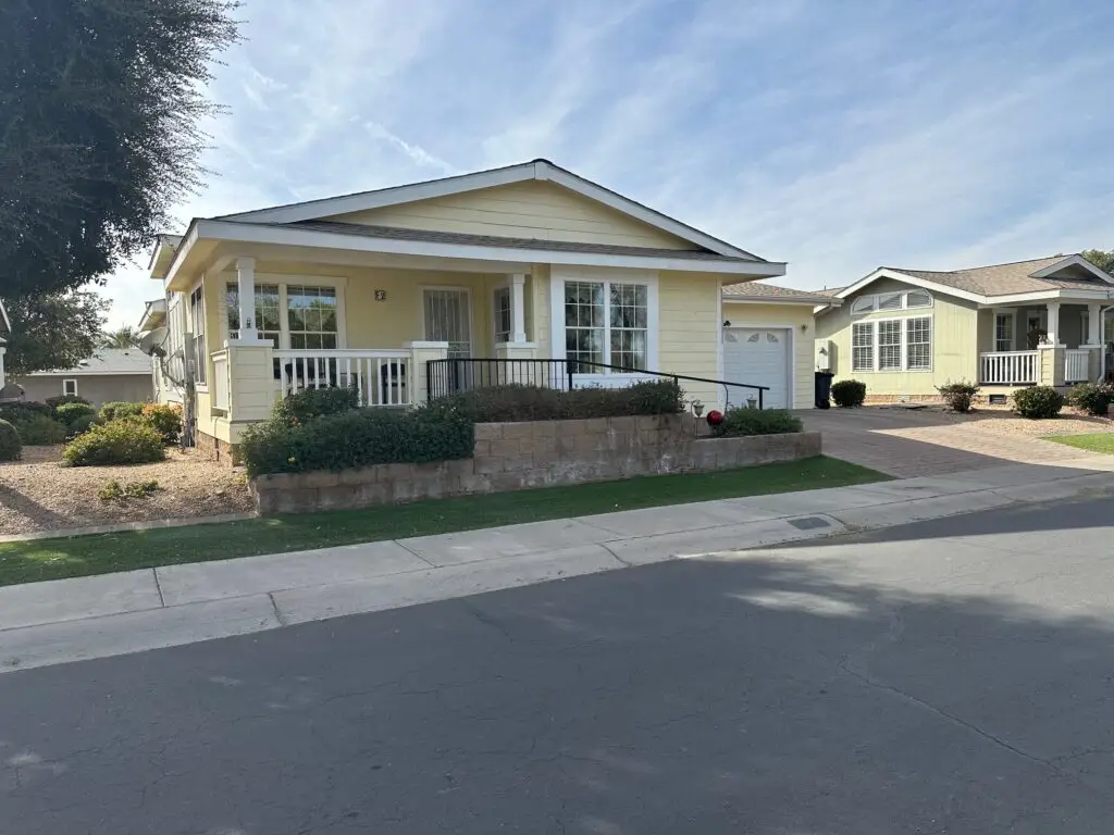 A single-story yellow house with a wraparound porch and white trim, set in a suburban neighborhood with a well-maintained garden and paved driveway.