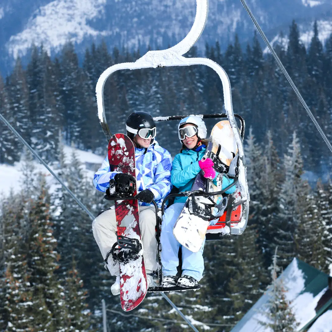 Two snowboarders on a ski lift, clad in winter gear and helmets, holding their snowboards against the backdrop of snowy trees, embark on thrilling adventures in Park City's winter skiing paradise.