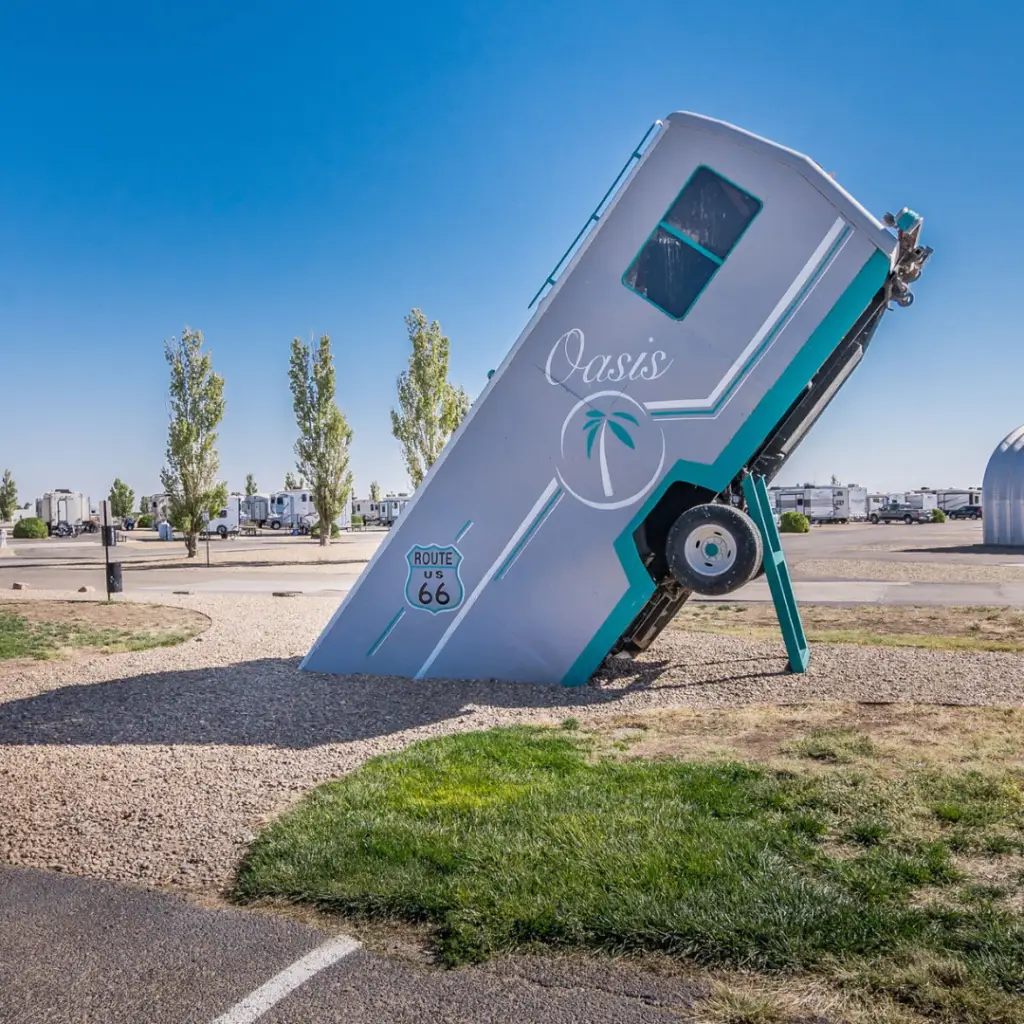 A vintage trailer, reminiscent of long-term RV travelers' adventures, is partially buried in the ground at an angle. Adorned with "Amarillo" and "Route 66," it rests under a clear blue sky.