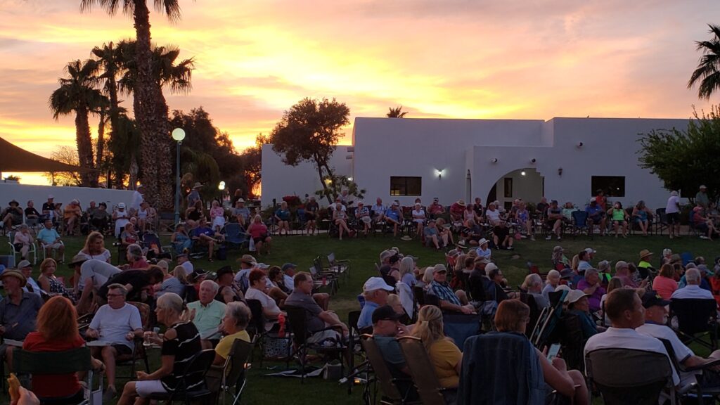 During a vibrant sunset in El Mirage, AZ, a large crowd sits on the grassy area, embarking on spring adventures. Modern white buildings and palm trees paint the backdrop of this picturesque scene.