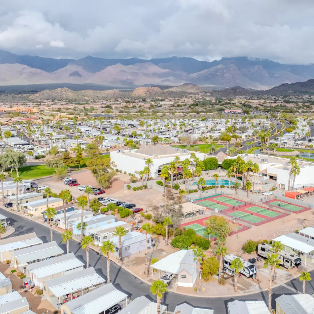 Aerial view of a residential community in Arizona with palm trees, tennis courts, and RVs set against a backdrop of mountains under a cloudy sky—perfect for winter visitors seeking an early booking.