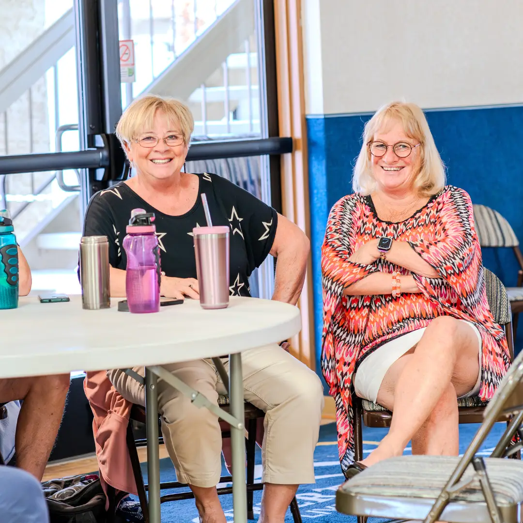 Two women sit at a round table indoors, smiling, as they enjoy activities at Sunrise RV Resort. The table holds pink and silver cups, capturing the spirit of January. A doorway is visible in the background.