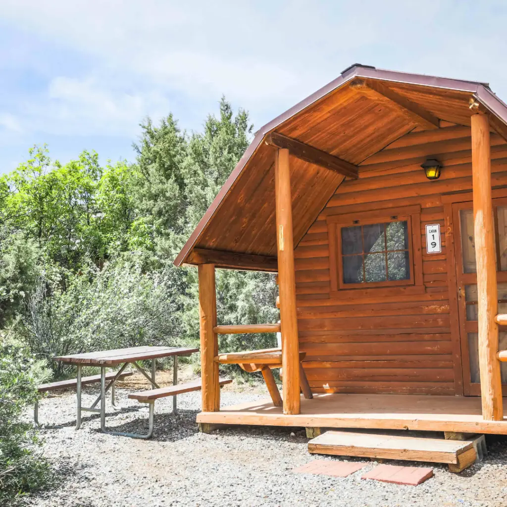 A small wooden cabin with a covered porch, lit by an outdoor bulb, stands next to a picnic table. It's the perfect Spring getaway, surrounded by trees and shrubs under a blue sky in Durango, Colorado.