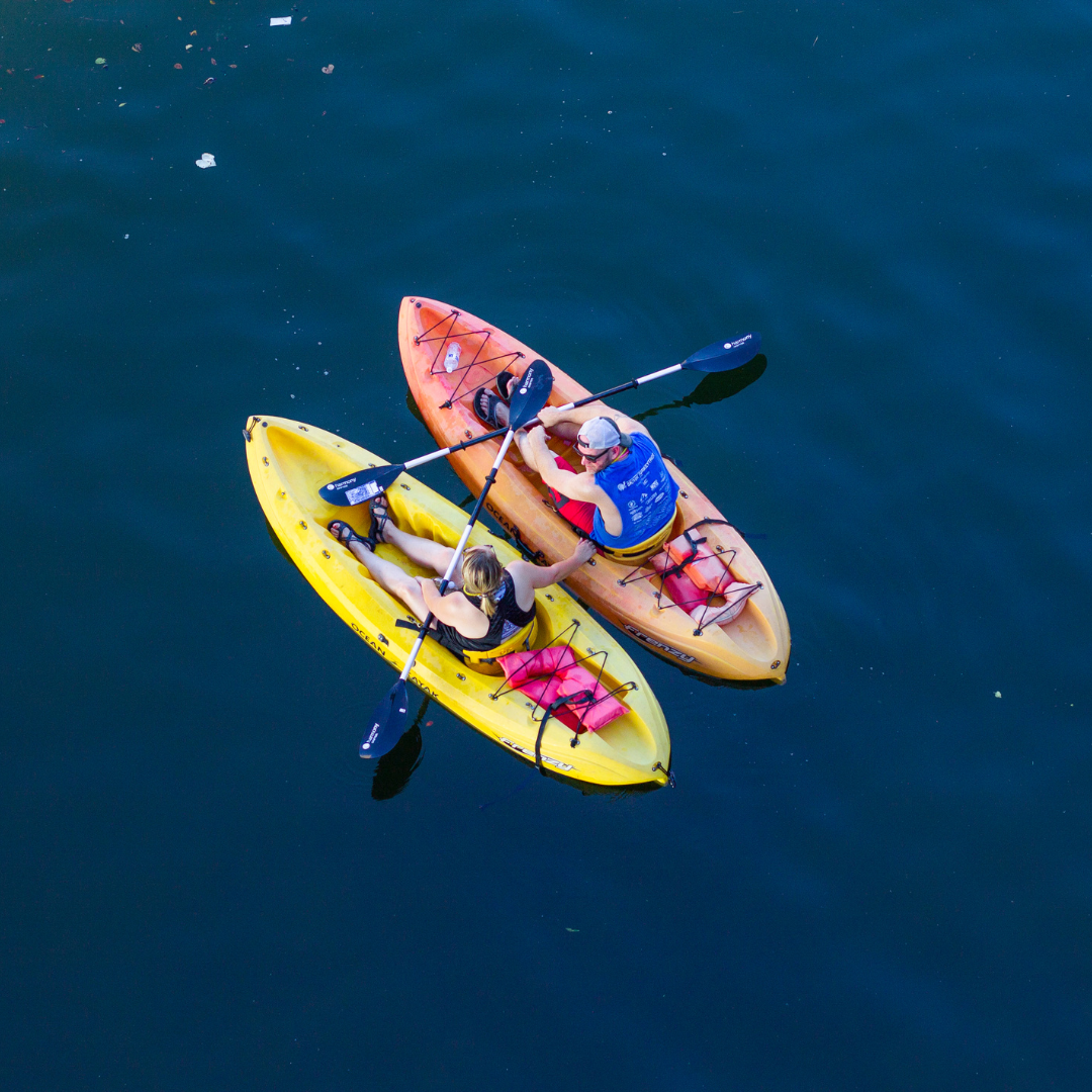 On a summer vacation, two people kayak on calm, dark blue water in yellow and orange kayaks, gently touching paddles.