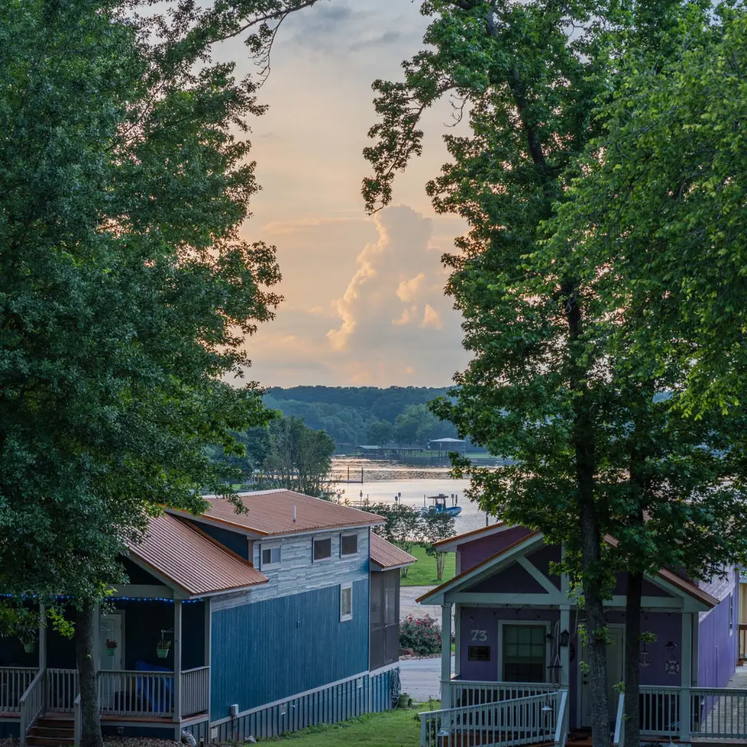 Cottages with colored roofs, perfect for those planning early for a spring on Lake Conroe, are nestled among trees and overlook a serene lake at sunset.