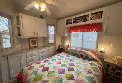 A small bedroom in Lot 53 features a colorful quilt on the bed. The room boasts white Cavco cabinets, a ceiling fan, and a window adorned with a red valance. A picture frame and lamp sit gracefully on the counter.