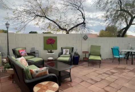 The outdoor patio at Lot 53 features green cushioned chairs, a glass-top table, and striking wall art of a red flower. Framed by trees and a white brick wall in the background, it's reminiscent of the charming design elements that defined Cavco's style back in 1995.