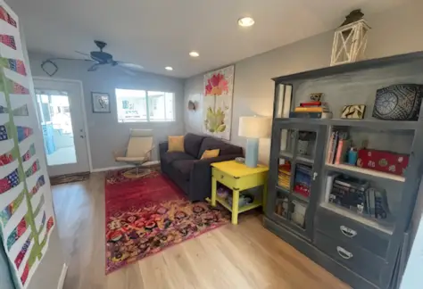 A living room with a dark sofa, vibrant rug, and a Cavco charm—featuring a yellow side table with a lamp. The gray cabinet, filled with books and decor from 1995, complements the white chair. Lot 53 includes a ceiling fan that completes this welcoming space.