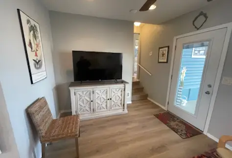 The living room in Lot 53 features a TV on a wooden stand, a woven chair to the left, and a rug on the floor. Cavco's craftsmanship is evident in the door leading outside. Walls are adorned with a framed picture and decorative items.