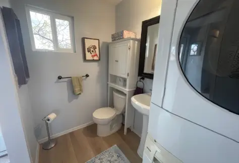 Compact bathroom featuring a toilet, sink, mirror, and storage cabinet. A washing machine sits snugly beside the window on the left. Adorned with a charming dog picture on the wall and a beige rug underfoot, this cozy space is reminiscent of Lot 53 in its efficient design.