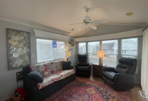 A cozy room in Lot 53 features a quilt-covered sofa, two recliners, a ceiling fan, and a rug. Lamps and large windows illuminate the space, while a decorative wall panel graces the left side, adding charm to this delightful Cavco home.