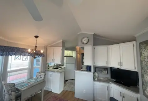The interior of Lot 53 features a small kitchen and dining area with white cabinets, a Cavco dining table, refrigerator, clock, and ceiling fan. The space boasts a cozy and compact layout.