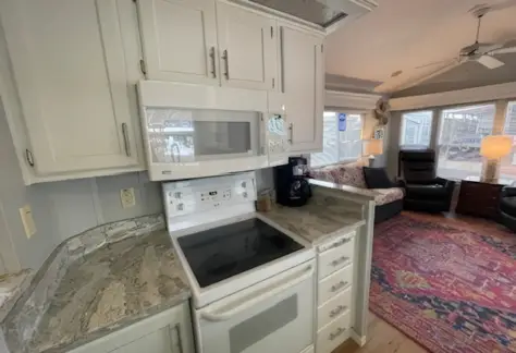 This modern kitchen in Lot 53 features white cabinets, an electric stove, and a microwave. The elegant marble countertop from the 1995 Cavco design extends into a cozy living area with large windows, a chic rug, and comfortable seating.