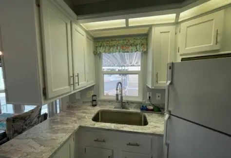 Nestled in Lot 53, this compact 1995 Cavco kitchen boasts white cabinets, a marble-patterned countertop, and a stainless steel sink. A refrigerator stands ready, while a window with a floral curtain graces the view above the sink.