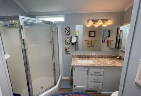 This Cavco-designed bathroom features a glass shower, a white vanity topped with marble, and wall-mounted lights. Small artworks adorn the walls, while a round, colorful rug from Lot 53 graces the floor, complementing the neatly arranged towels.