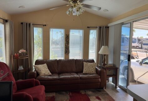Living room with a 1995 Fleetwood brown leather sofa, red recliner, and a floor lamp near sliding glass doors. Ceiling fan above, large windows with curtains, and a small table with flowers to the left. Lot 171 offers charming comfort and style in every corner.