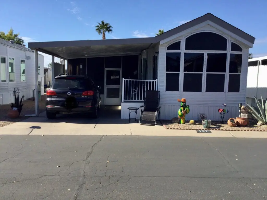 A charming 1998 Chariot Eagle mobile home with a car parked in the driveway. The yard boasts a whimsical cactus sculpture and potted plants, while a sleek black lounge chair rests invitingly under the awning.